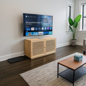 Una sala de estar con un televisor de pantalla plana montado en la pared, un mueble de madera debajo, un tapete negro en el suelo, una mesa de centro con una caja de pañuelos y una planta en maceta.