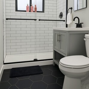 Ein Badezimmer mit einer weißen Toilette, einer schwarzen Matte, einem Fenster mit Toilettenartikeln und einem Handtuchhalter.