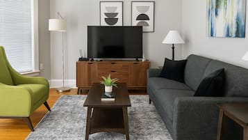 A living room with a flat-screen TV, a green armchair, a grey sofa, and a small table with a plant.
