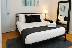 A neatly made bed with a black frame, white bedding, and two black pillows. There is a bedside table with a lamp and a folded towel. A large mirror is on the wall to the right. - 2BR Steps from Wrigley Field (Chicago)