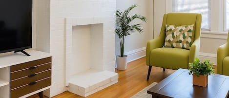 Un salon avec une cheminée en briques blanches, un téléviseur posé sur un meuble en bois, un fauteuil vert et une table basse.