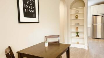 Een eetgedeelte met een houten tafel en stoelen, een kleine potplant en een wandplank met decoratieve objecten.