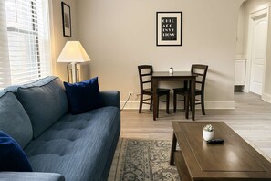 A living room with a blue sofa, a coffee table, and a dining table with chairs. There is a lamp on the sofa and a framed picture on the wall. - Portage Park 1BR Retreat (Chicago)