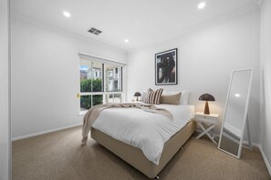 A bedroom with a large bed, a bedside table with a lamp, a window, and a framed picture on the wall. - Burleigh: Easy Coastal Living on Broadway (Glenelg South)