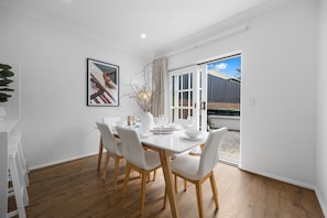 A dining area with a white table set for four, wooden chairs, a framed picture on the wall, and a view through a glass door to the outside. - Burleigh: Easy Coastal Living on Broadway (Glenelg South)