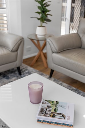 A living room with two leather sofas, a glass coffee table, a purple candle, and a magazine. - Burleigh: Easy Coastal Living on Broadway (Glenelg South)