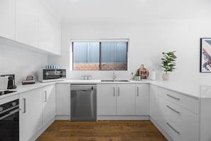 A compact kitchen with white cabinetry, a dishwasher, a microwave, and a sink. - Burleigh: Easy Coastal Living on Broadway (Glenelg South)