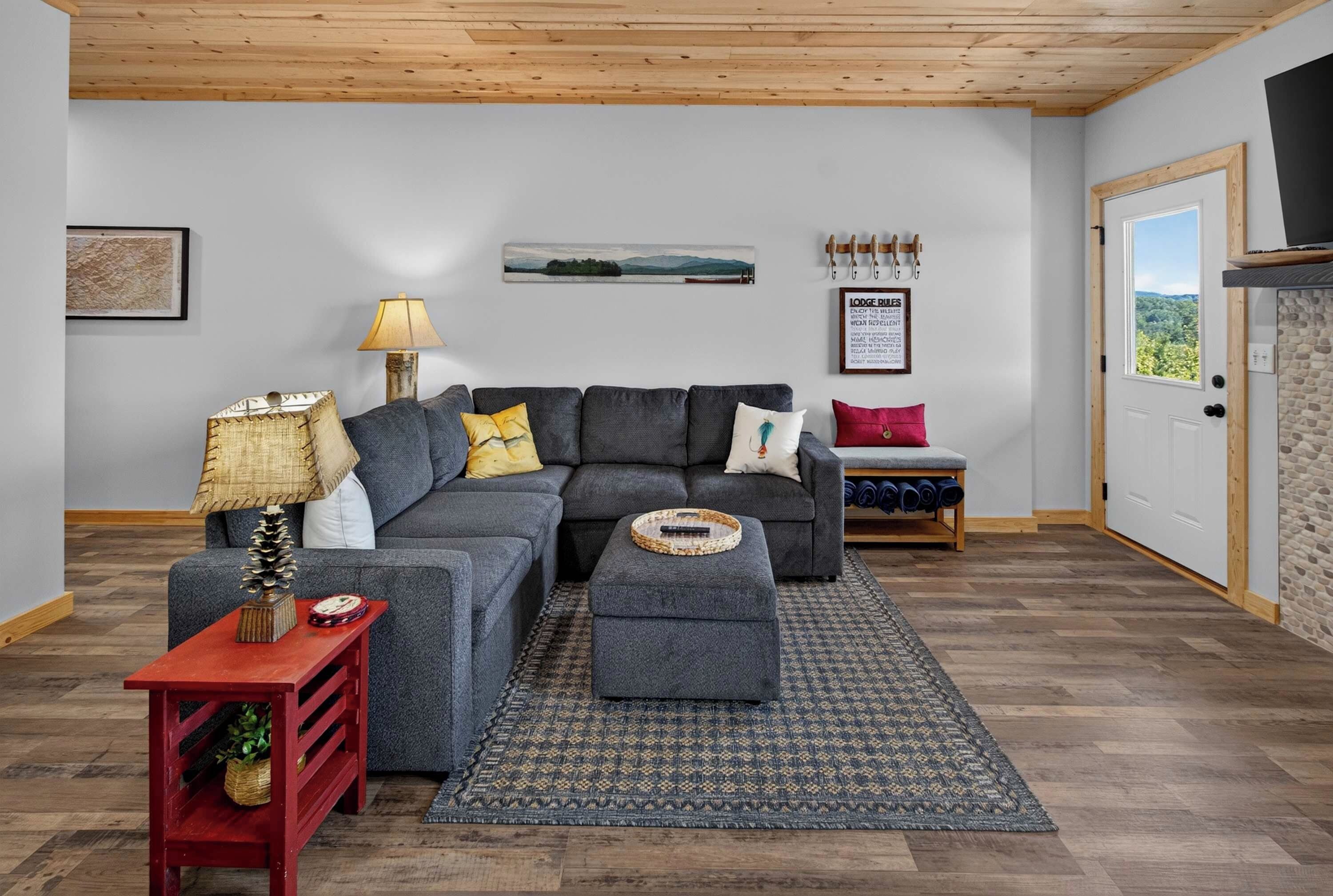 A living room with a sectional sofa, a red side table with a lamp, a patterned rug, and a door leading to a scenic view.