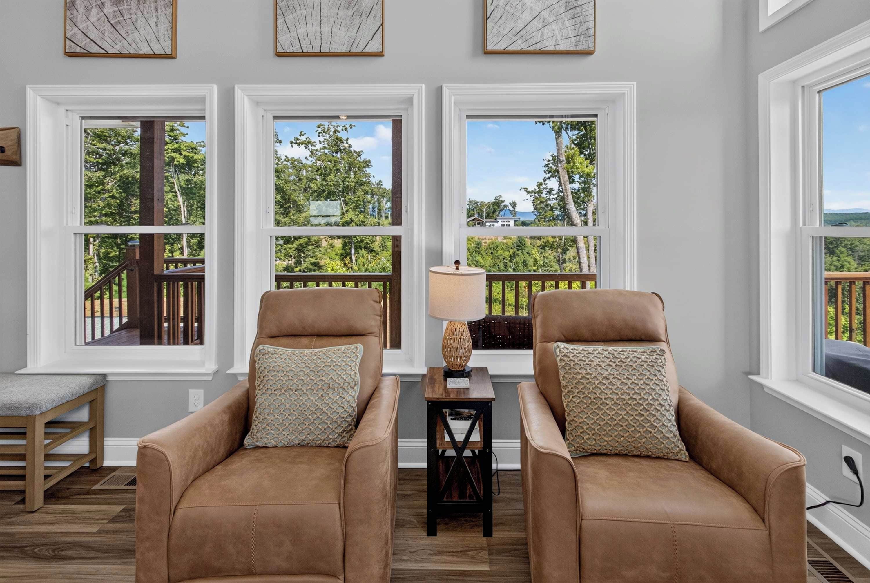 A room with two leather armchairs, a small table with a lamp, and a view of a deck and trees through large windows.