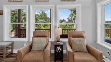 A room with two leather armchairs, a small table with a lamp, and a view of a deck and trees through large windows.