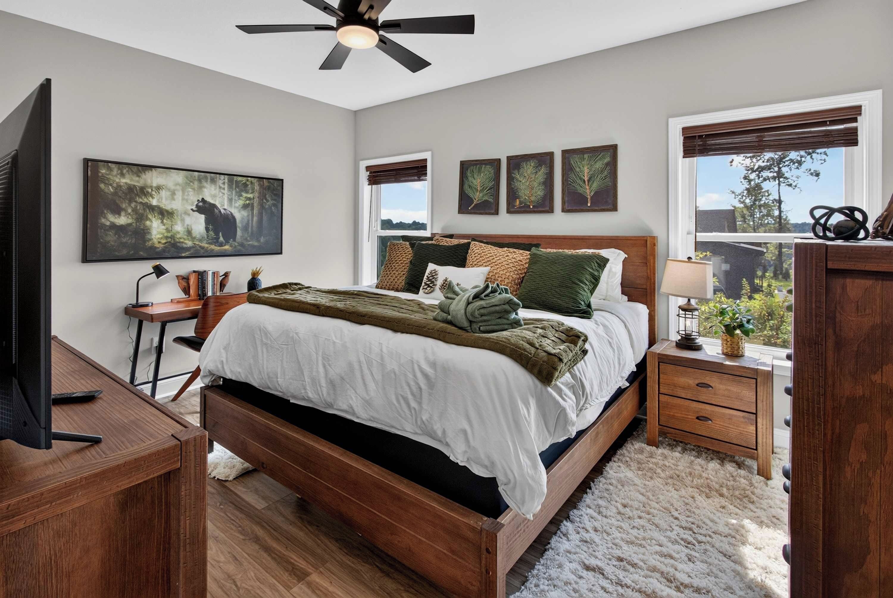 A bedroom with a bed, a desk, a chair, a television, and a window with blinds.