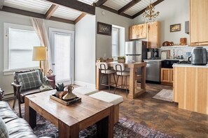A cozy living room with a wooden table, a sofa, and a kitchen area with wooden cabinets and a refrigerator. - Walk to Greek Peak Lifts - Mtn View & Balcony (Cortland)