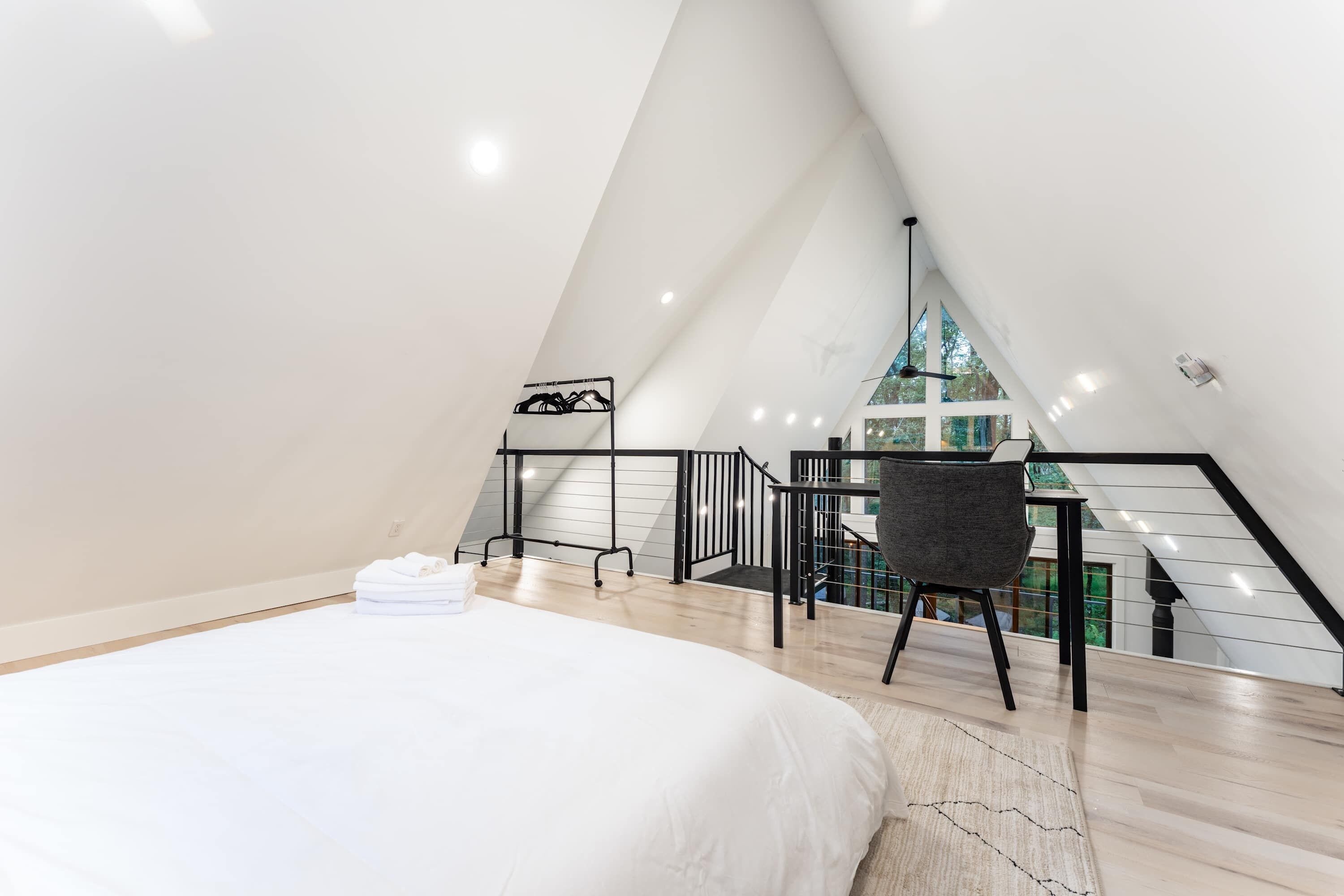 A modern loft bedroom with a bed, a chair, and a view of a green landscape through large windows.