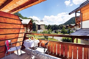 Ein hölzerner Balkon mit Tisch und Stühlen, der auf eine gebirgige Landschaft mit Gebäuden blickt.