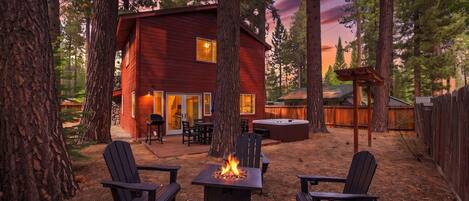 A cabin with a fire pit and outdoor seating, surrounded by tall trees.