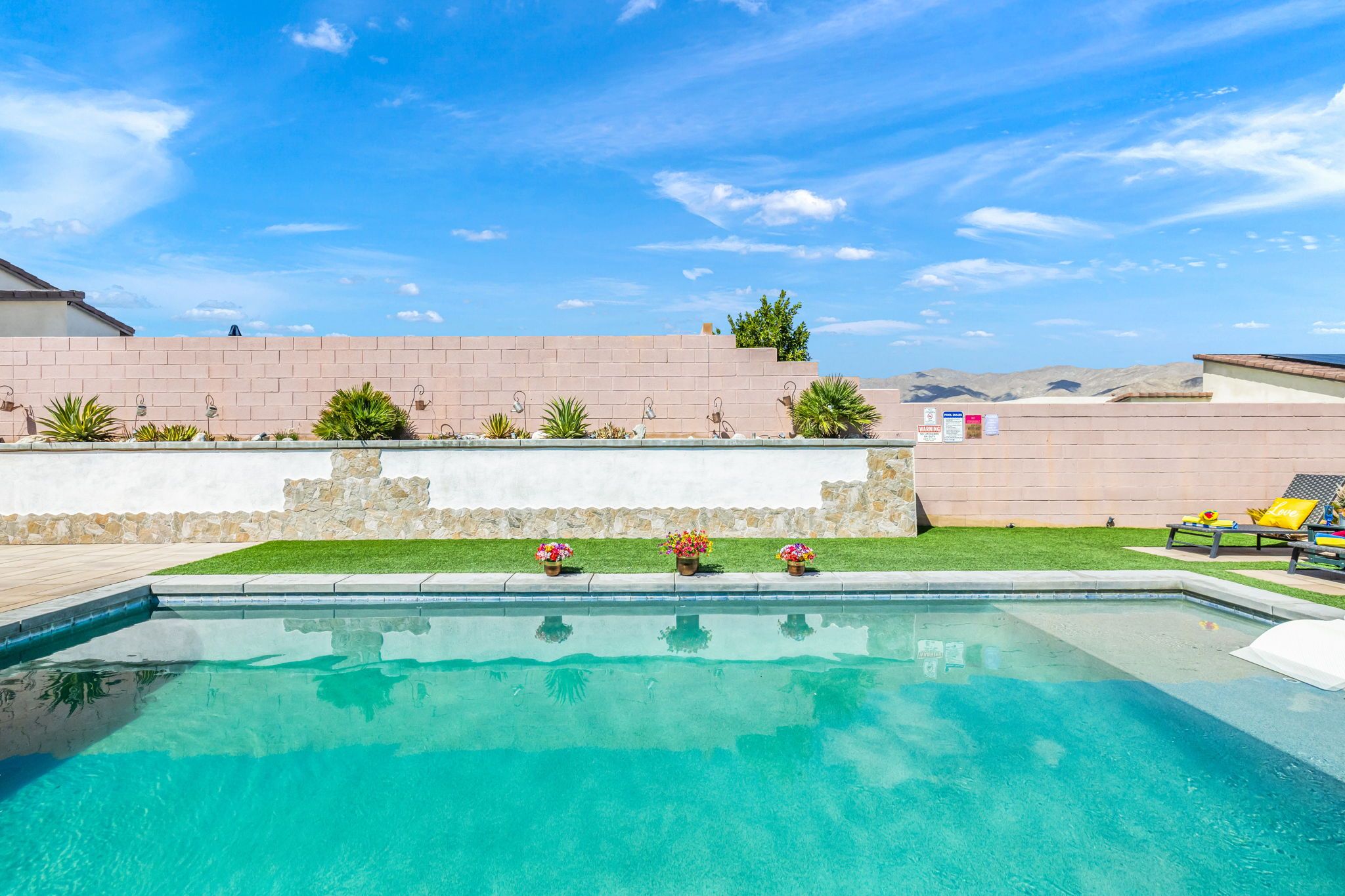 A clear swimming pool with a stone wall and a grassy area with potted plants.