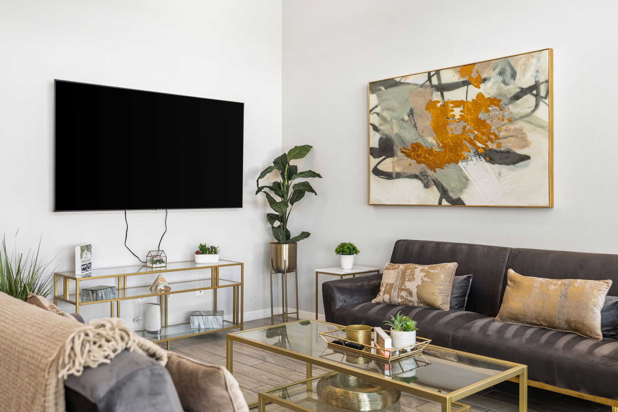A modern living room with a flat-screen TV, a dark sofa, a glass coffee table, and a large abstract painting.