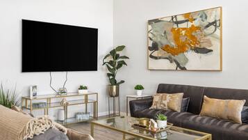 A modern living room with a flat-screen TV, a dark sofa, a glass coffee table, and a large abstract painting.