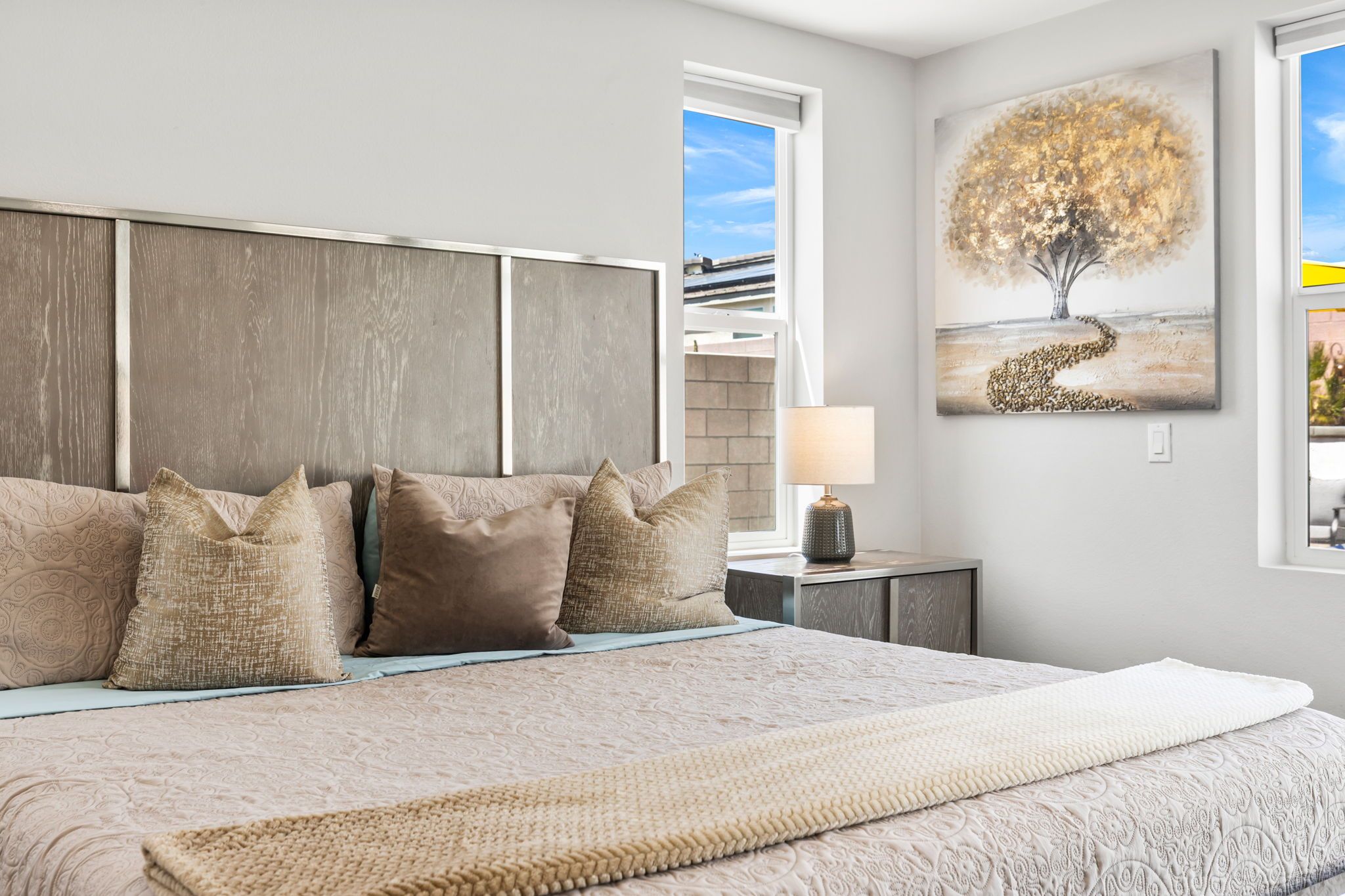 A bedroom with a large bed, a headboard, a nightstand with a lamp, and a painting of a tree.