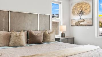 A bedroom with a large bed, a headboard, a nightstand with a lamp, and a painting of a tree.