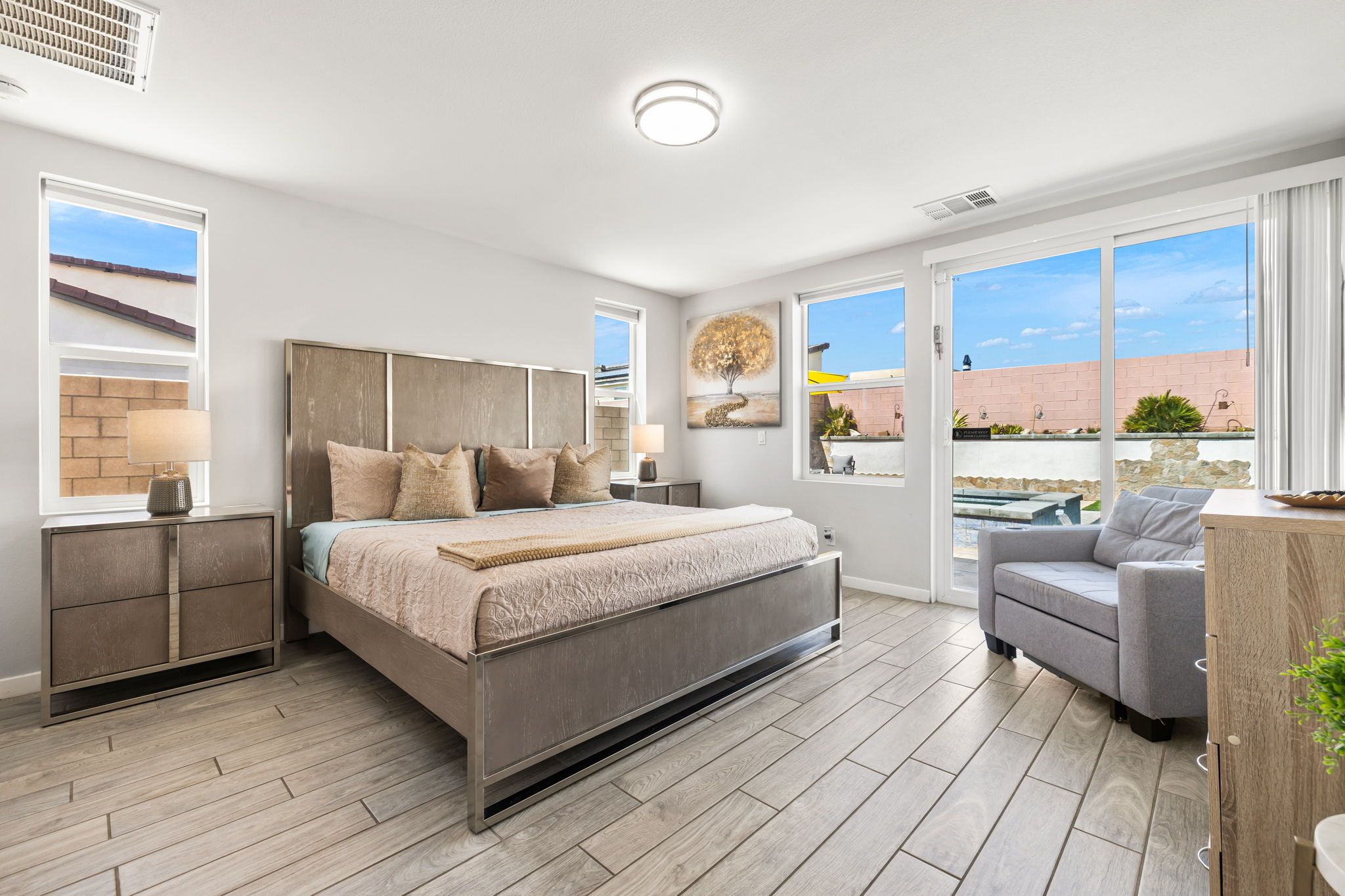 A modern bedroom with a bed, bedside tables, a grey sofa, and large windows offering a view of the outdoors.