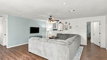 A living room with a grey sofa, a flat-screen TV, a ceiling fan, and a wooden floor.