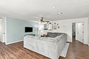 A living room with a grey sofa, a flat-screen TV, a ceiling fan, and a wooden floor.