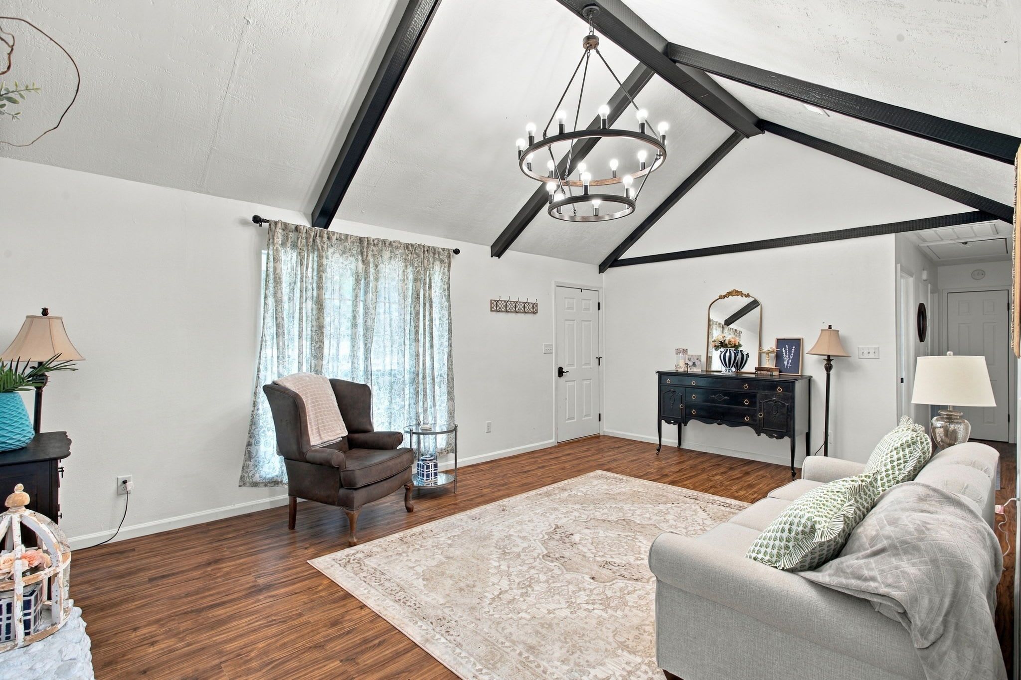 A room with a high ceiling, exposed beams, a chandelier, a brown leather chair, a grey sofa, a wooden floor, and a patterned rug.