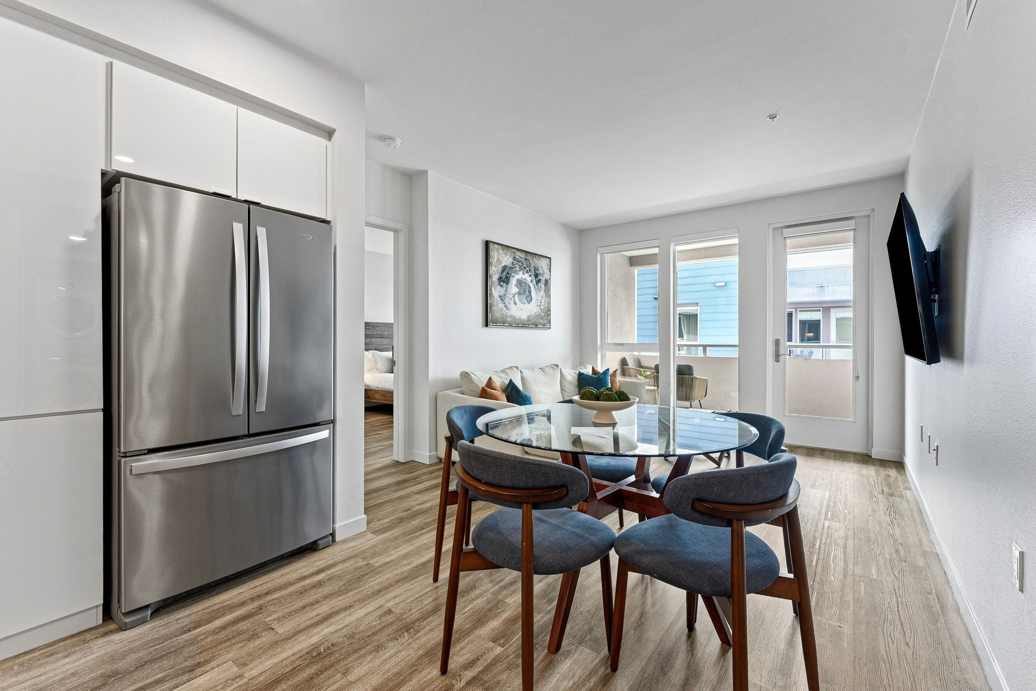 A modern kitchen with a stainless steel refrigerator, a glass-top dining table with four chairs, and a view of the outside through large sliding glass doors.