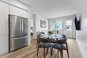 A modern kitchen with a stainless steel refrigerator, a glass-top dining table with four chairs, and a view of the outside through large sliding glass doors.