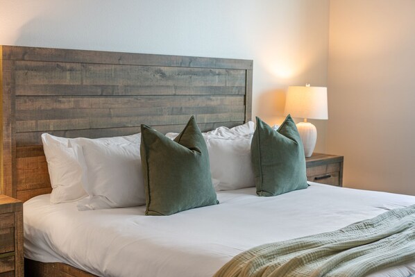 A neatly made bed with white linens and two green pillows, a wooden headboard, a bedside lamp, and a bedside table.