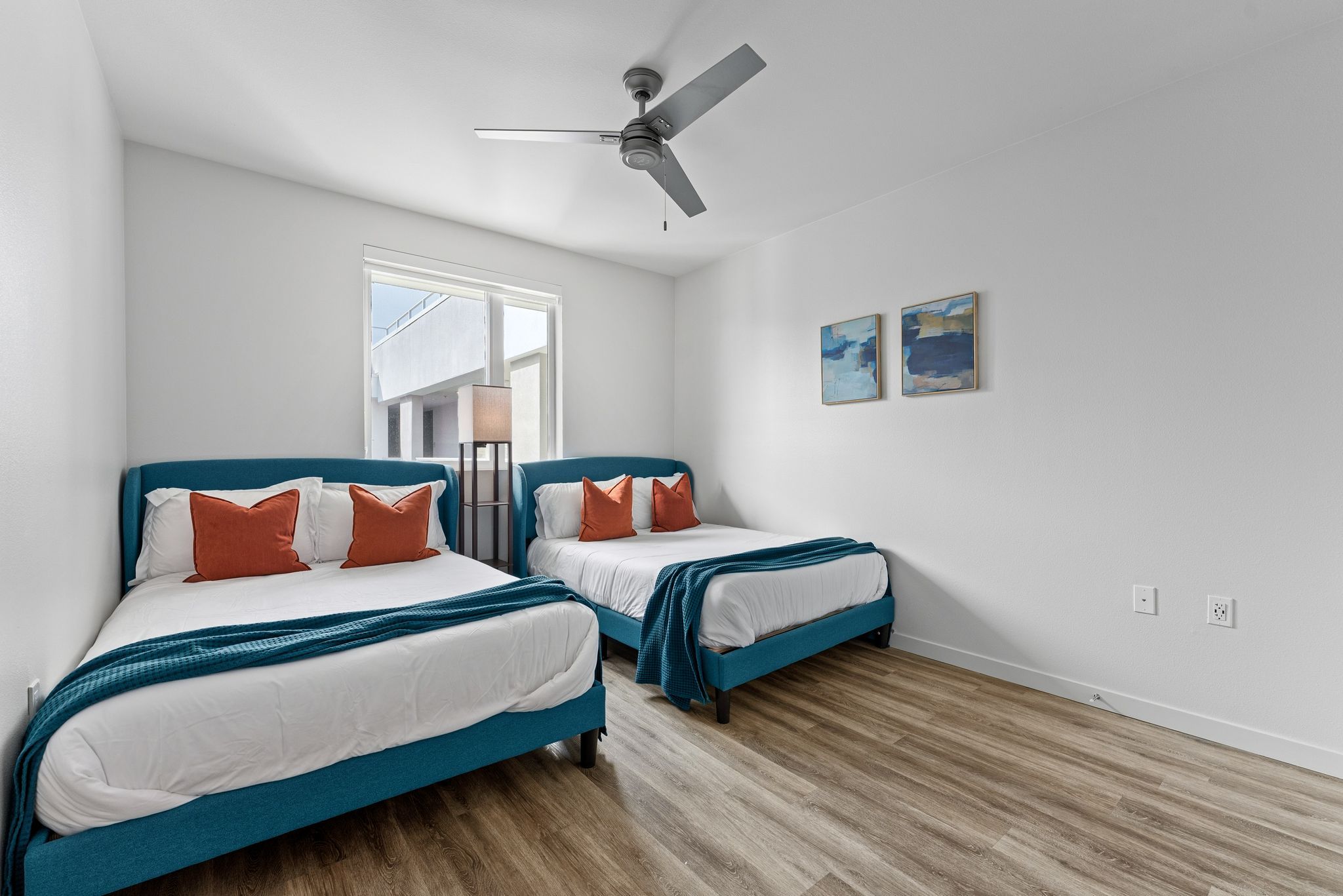A bedroom with two beds, a ceiling fan, a window, and two wall-mounted artworks.