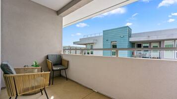 A balcony with a wicker chair and a small table, overlooking a modern building with a blue facade.