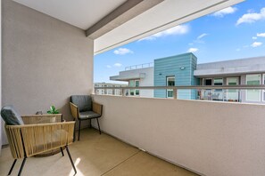 A balcony with a wicker chair and a small table, overlooking a modern building with a blue facade.