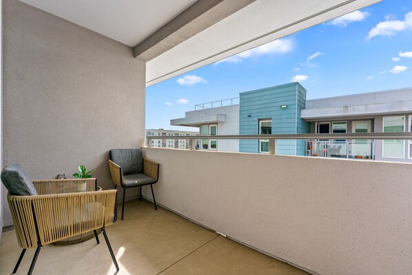 A balcony with a wicker chair and a small table, overlooking a modern building with a blue facade.