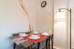 Ein Esstisch ist mit Tellern, Gläsern und Besteck gedeckt, an der Wand hängt eine Uhr und eine Lampe steht daneben.