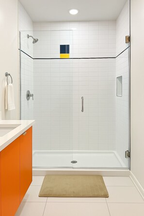 A modern bathroom with a glass-enclosed shower, white tiled walls, and a beige bath mat. - Elegant 2BR Apartment w/ In-Unit Laundry (Chicago)