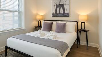 A hotel room with a bed, two bedside lamps, a window with blinds, and a wall-mounted picture of a bridge.