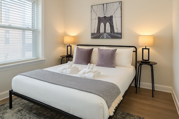 A hotel room with a bed, two bedside lamps, a window with blinds, and a wall-mounted picture of a bridge. - Cozy 2BR Apt for a Seamless Stay (Chicago)