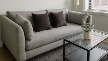 A modern living room with a grey sofa, two dark cushions, a glass coffee table, and a potted plant.