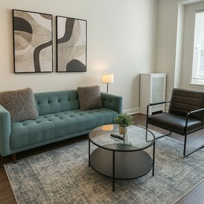 Een woonkamer met een blauwgroene bank, een zwarte lederen fauteuil, een glazen salontafel en abstracte wanddecoratie.