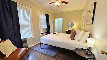 A bedroom with a bed, a chair, a table, a ceiling fan, and a window with blinds.