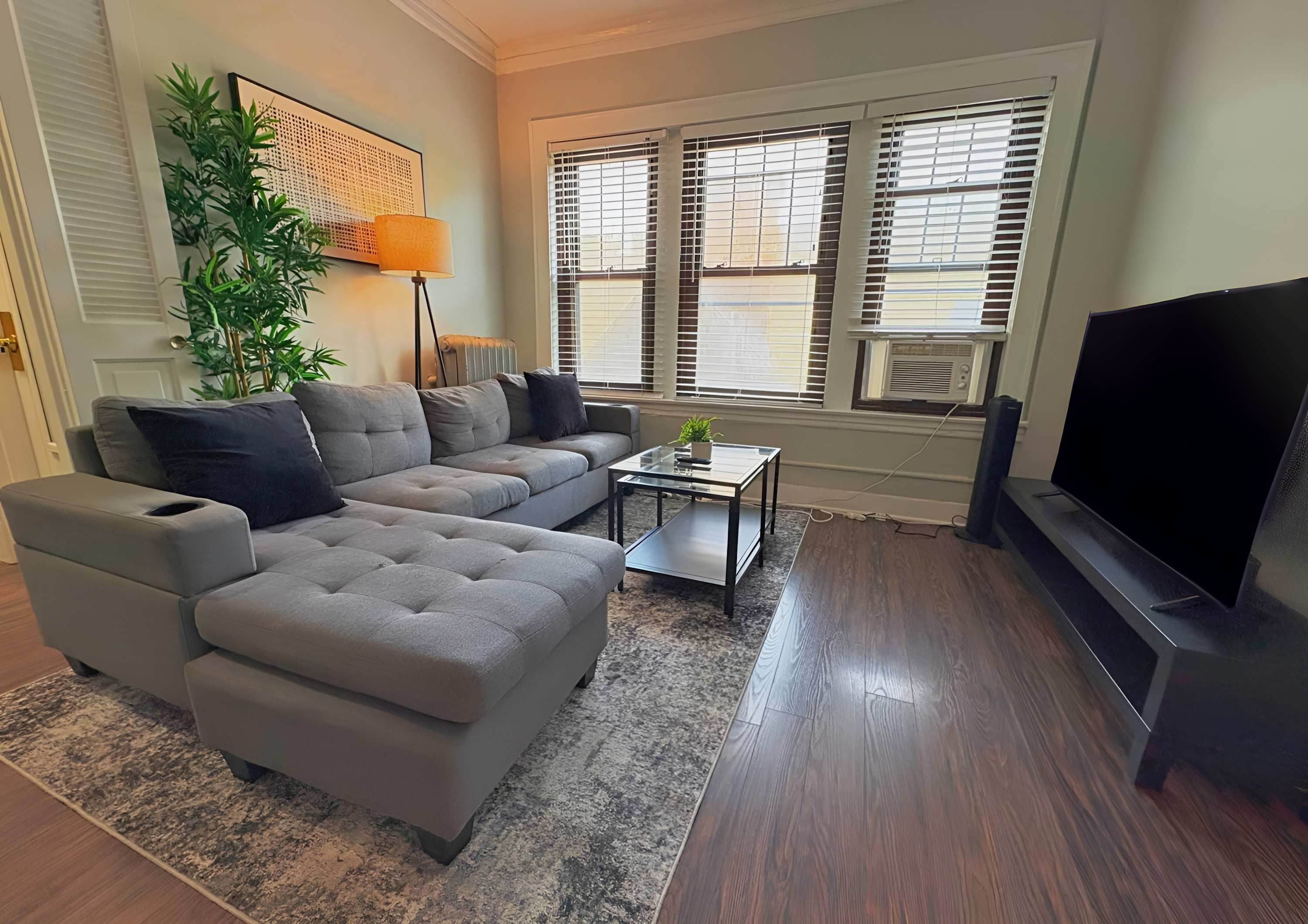 A living room with a sectional sofa, a coffee table, a flat-screen TV, and a large window with blinds.