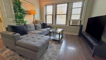 A living room with a sectional sofa, a coffee table, a flat-screen TV, and a large window with blinds.