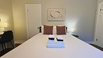 A hotel room with a large bed, two pillows, a bedside table with a lamp, and a door.