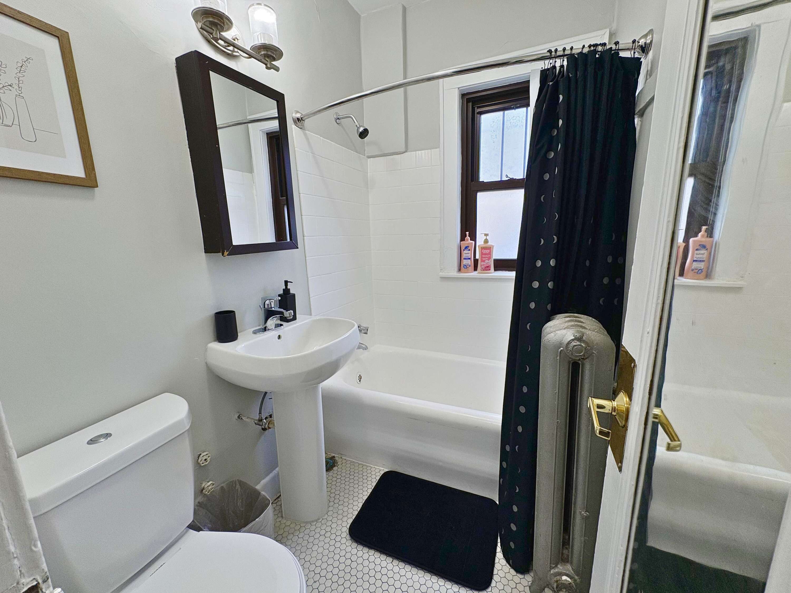 A bathroom with a white sink, a toilet, a bathtub, and a shower. There is a window with a curtain, and toiletries are visible on the windowsill.