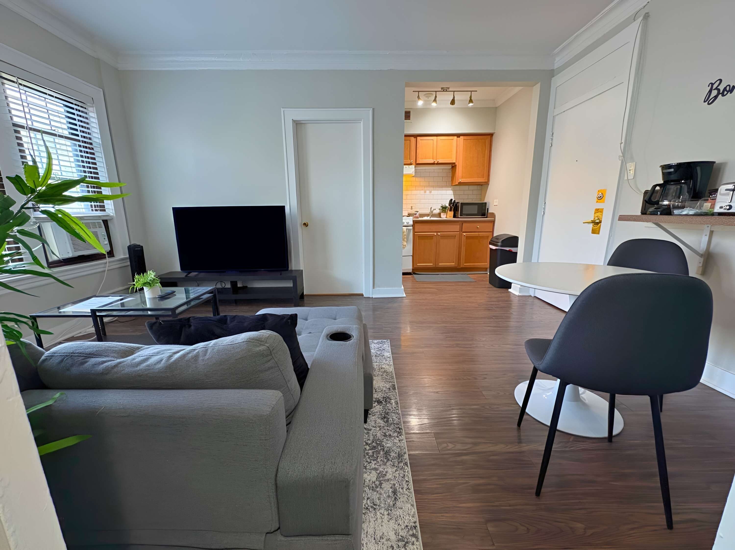 A modern living room with a grey sofa, a flat-screen TV, and a coffee table. A kitchen is visible through an open door, featuring wooden cabinets and a microwave.