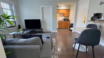 A modern living room with a grey sofa, a flat-screen TV, and a coffee table. A kitchen is visible through an open door, featuring wooden cabinets and a microwave.