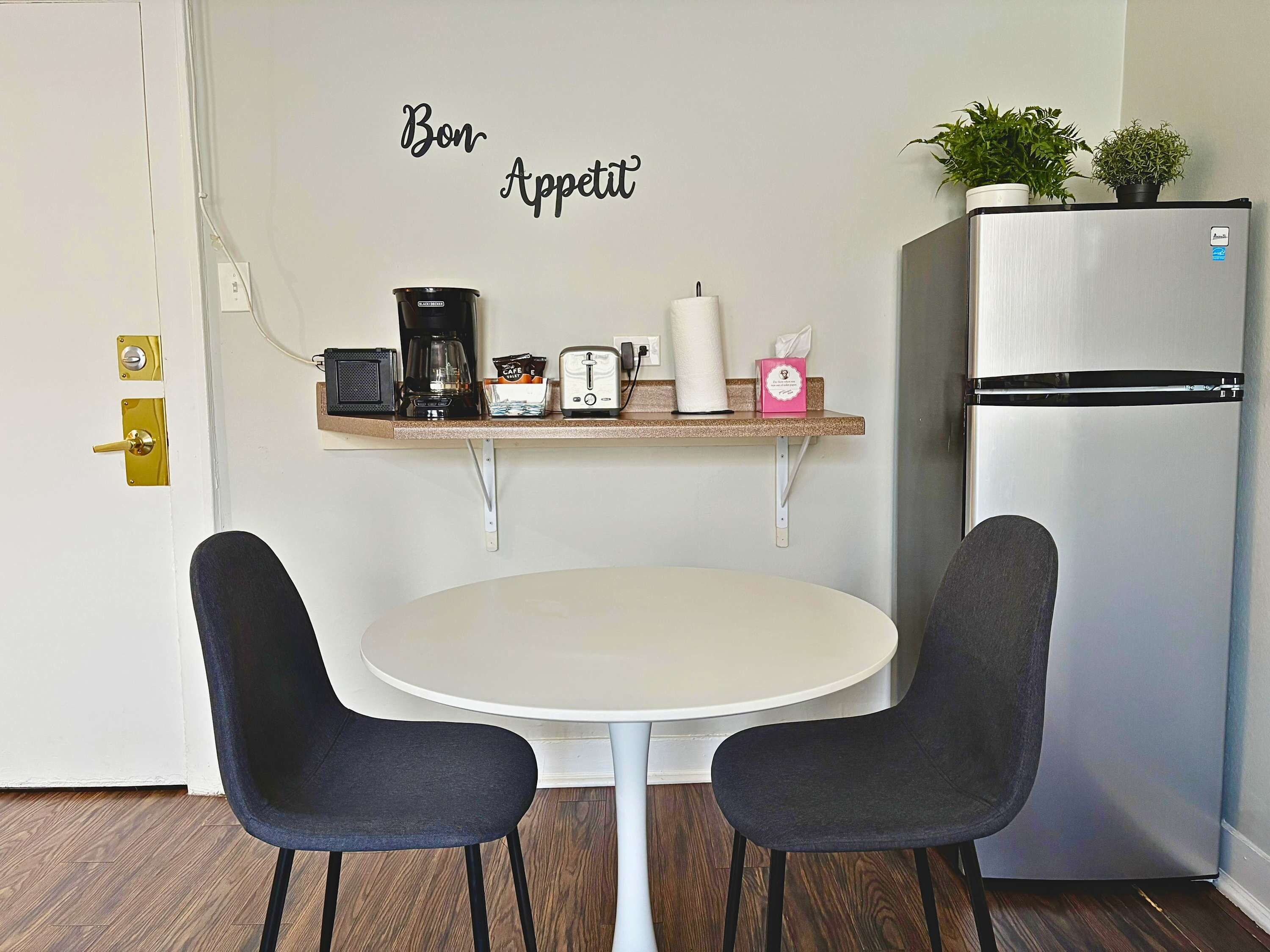 A small kitchenette with a white refrigerator, a coffee maker, a toaster, and a shelf with various items.