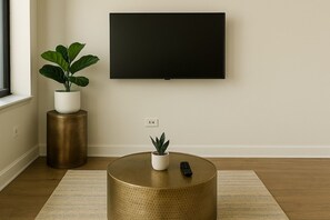 Un salon moderne avec un téléviseur à écran plat fixé au mur, une table ronde dorée ornée d’une plante en pot et une télécommande posée sur la table.